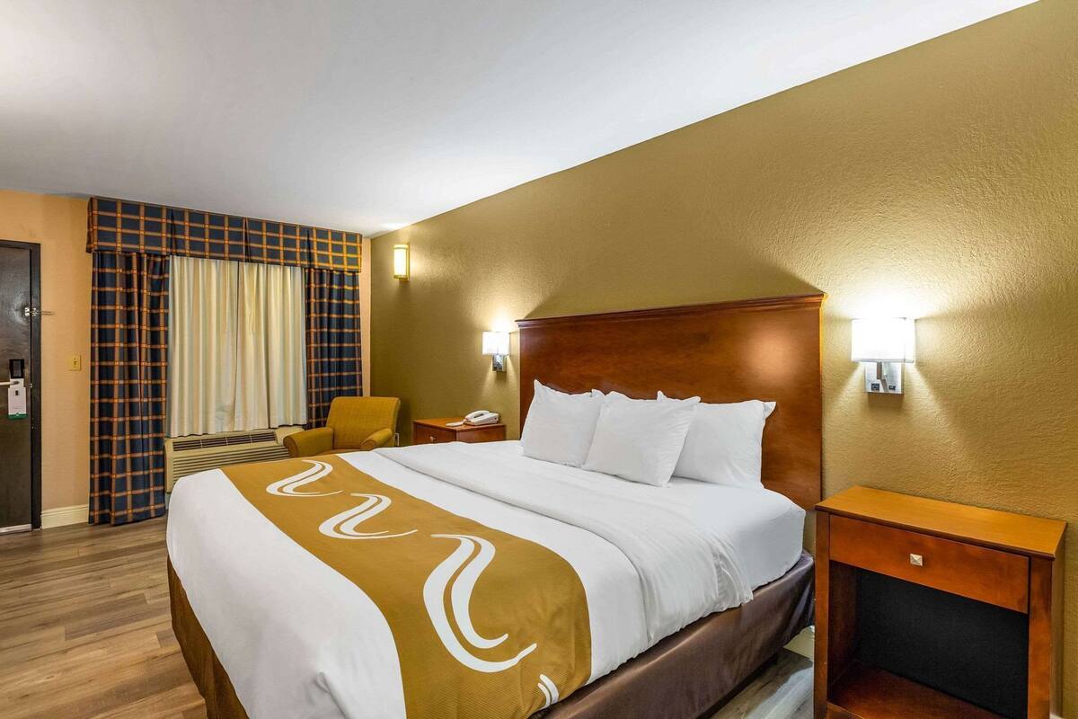Quarto de hotel com cama, uma cadeira, mesa de cabeceira e luminária fixada na parede.