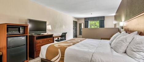 A hotel room with a bed, a desk, a television, a refrigerator, and a window with a view of greenery.