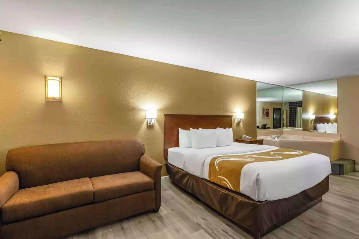 Quarto de hotel com uma cama grande, um sofá marrom, um banheiro com banheira e um espelho refletindo o quarto.