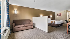 Habitación de hotel con un sofá marrón, una cama con cabecera blanca, un escritorio y una lámpara fijada a la pared.