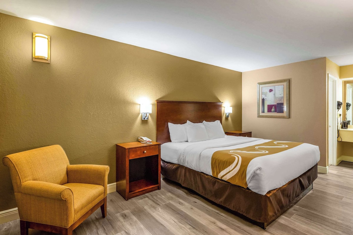 Quarto de hotel com cama, uma cadeira, mesa de cabeceira e luminária na parede.