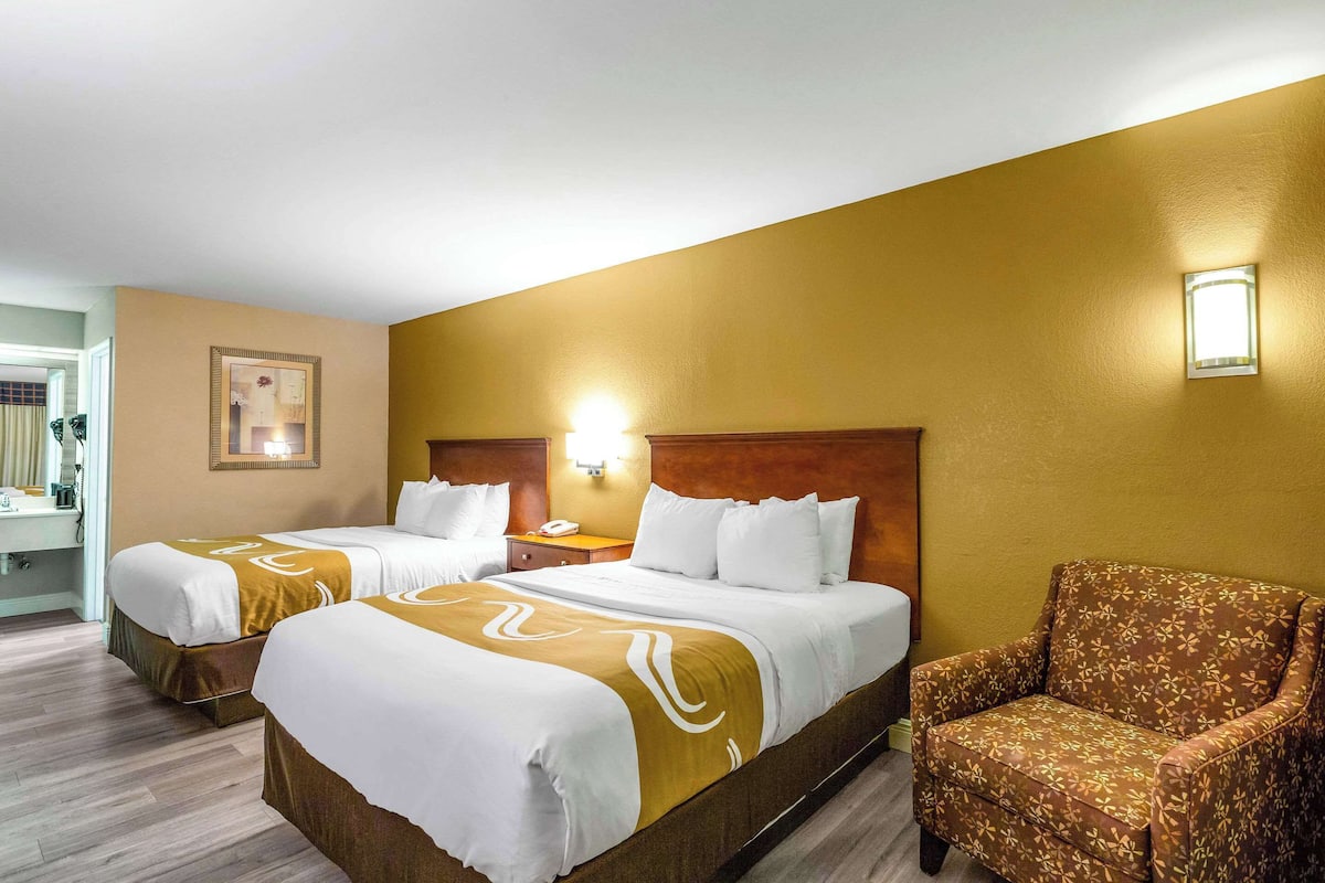 Quarto de hotel com duas camas, uma cadeira, uma mesinha de cabeceira e uma luminária fixada na parede.