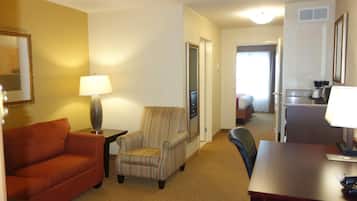 A hotel room with a sofa, armchair, desk, lamp, and a kitchenette area.
