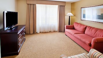 A hotel room with a TV on a dresser, a red sofa, a striped chair, a lamp, and a large window with curtains.