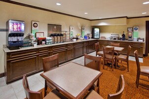 Un área de desayuno de hotel con una estación de bebidas de autoservicio, una barra de buffet y arreglos de asientos.