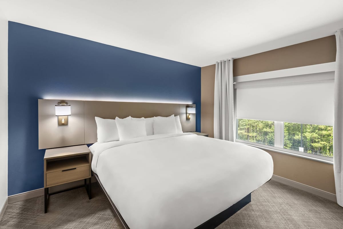 Quarto de hotel com uma cama grande, mesa de cabeceira, luminária na parede e janela com cortinas.