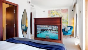 Une chambre d’hôtel avec un lit, un téléviseur diffusant des images d’une piscine, un fauteuil bleu et une peinture abstraite aux couleurs vives accrochée au mur.