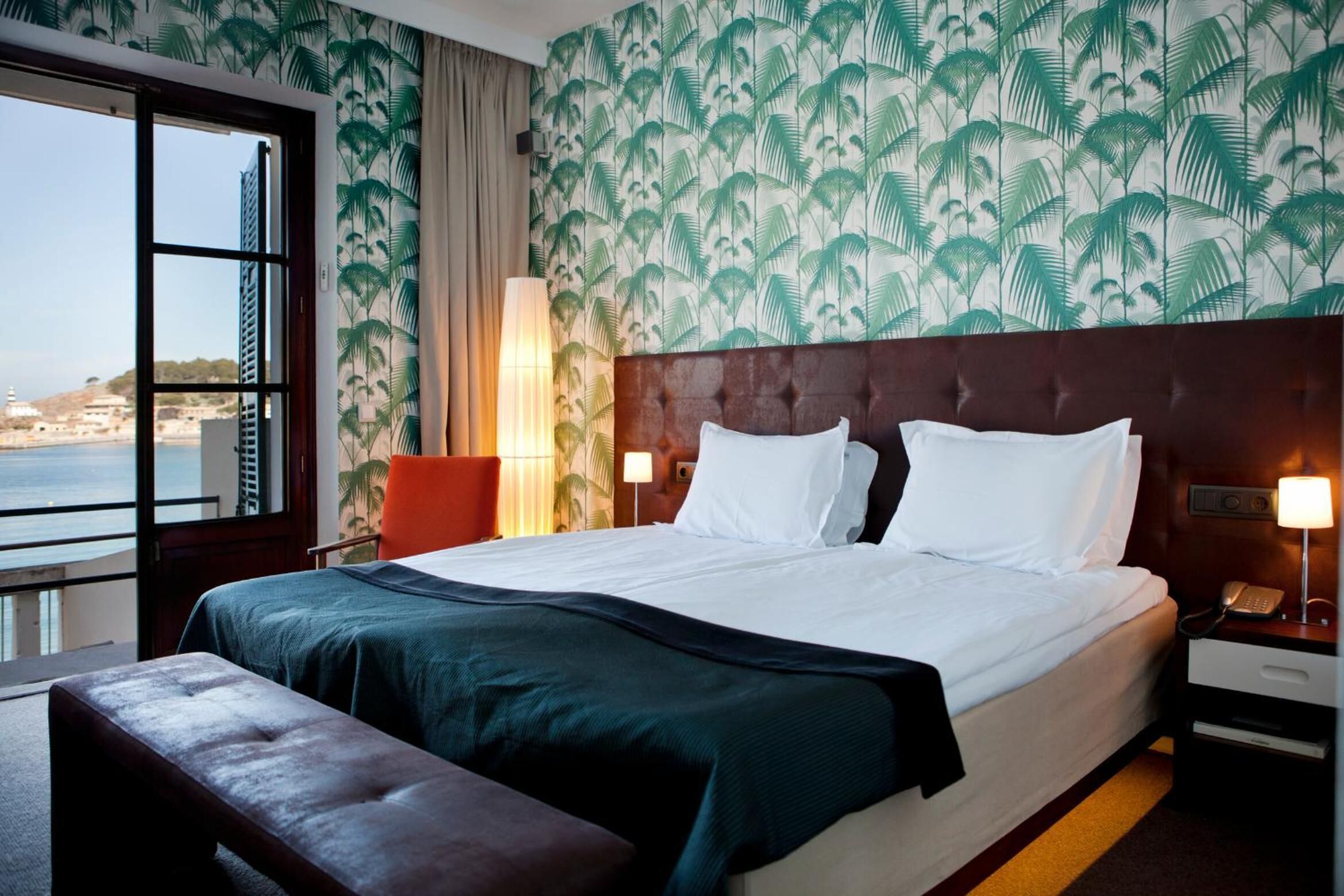 Quarto de hotel com uma cama grande, um banco, uma mesa de cabeceira e vista para o mar.