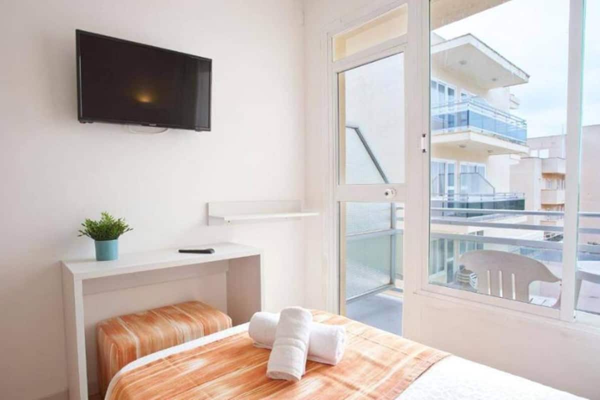 Een hotelkamer met een bed, een aan de muur bevestigde televisie, een klein nachtkastje met een plant en uitzicht op een balkon met andere gebouwen.