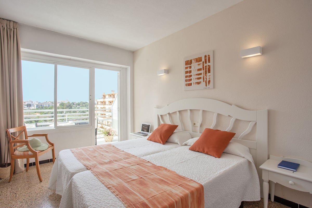 Een hotelkamer met twee bedden, een bureau, een stoel en een groot raam met uitzicht op de stad.