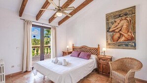 A bedroom with a bed, a ceiling fan, a painting, a wicker chair, and a balcony with curtains. - Villa Karen (Sa Rapita)