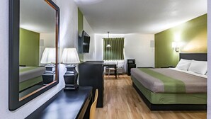 Et hotelværelse med en seng, et skrivebord med stol, et spejl, et fjernsyn og et vindue med gardiner.