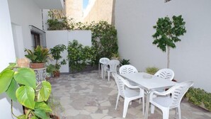 Een patio met een stenen vloer, witte plastic stoelen en een tafel. Er staan potplanten en een kleine boom.