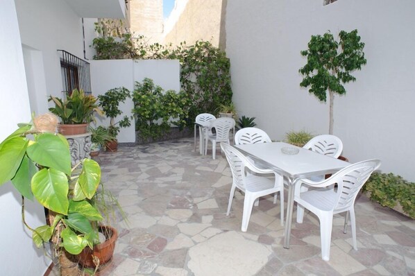 A patio area with a stone floor, white plastic chairs, and a table. There are potted plants and a small tree. - Montaña (San Antonio)