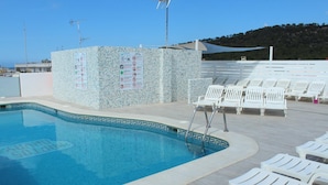 A swimming pool with a tiled edge, white lounge chairs, and a sign with various symbols and text. - Orvay Apartments (Sant Antoni de Portmany)