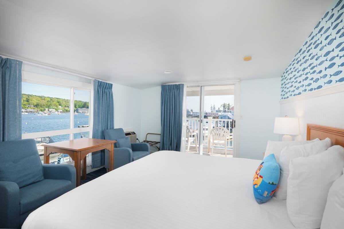 A hotel room with a large bed, a desk, two chairs, a view of the sea, and a balcony with a railing.