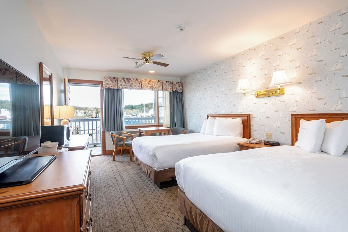 A hotel room with two beds, a desk, a chair, a ceiling fan, and a view of the harbor.