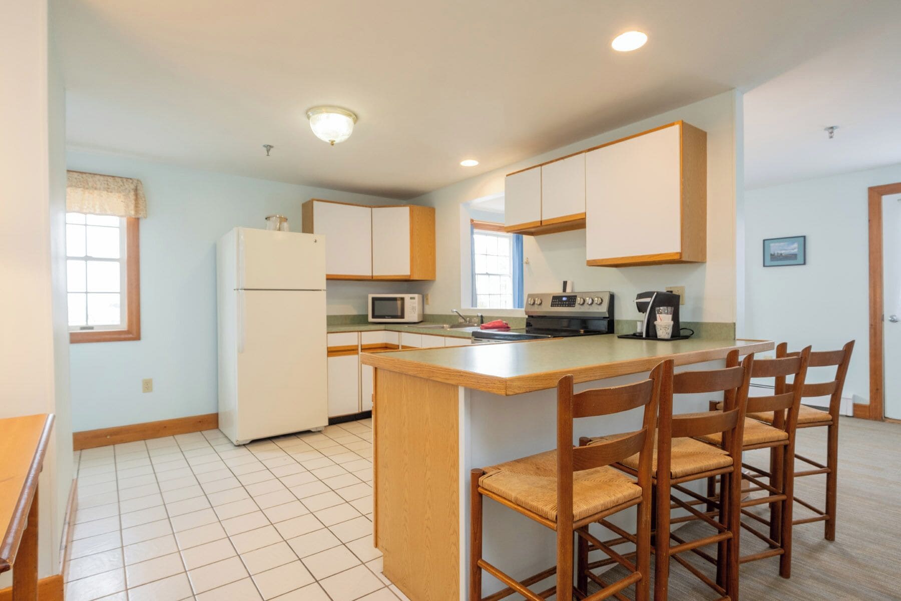 A compact kitchen with a central island, wooden chairs, a refrigerator, microwave, stove, and a coffee maker.
