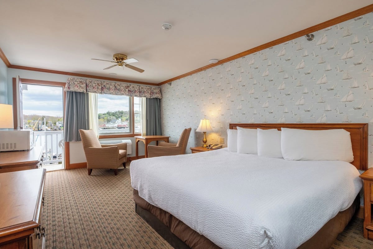 A hotel room with a large bed, two chairs, a desk, a ceiling fan, and a view of the outside.