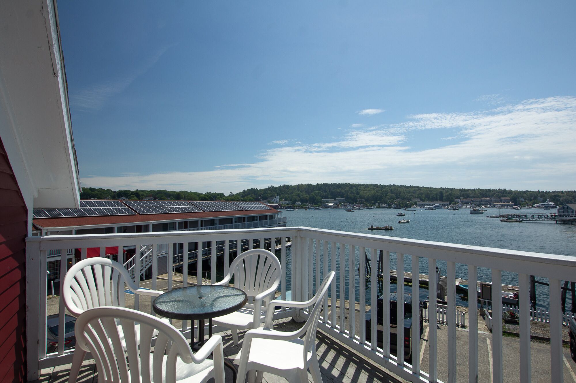 A balcony with white chairs and a small table, overlooking a marina with boats and a forested hill in the background.