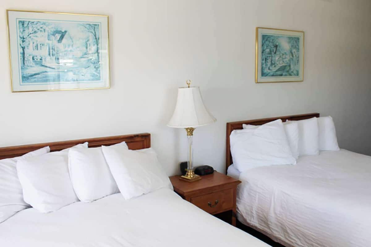 A hotel room with two beds, a nightstand with a lamp, and two framed artworks on the wall.
