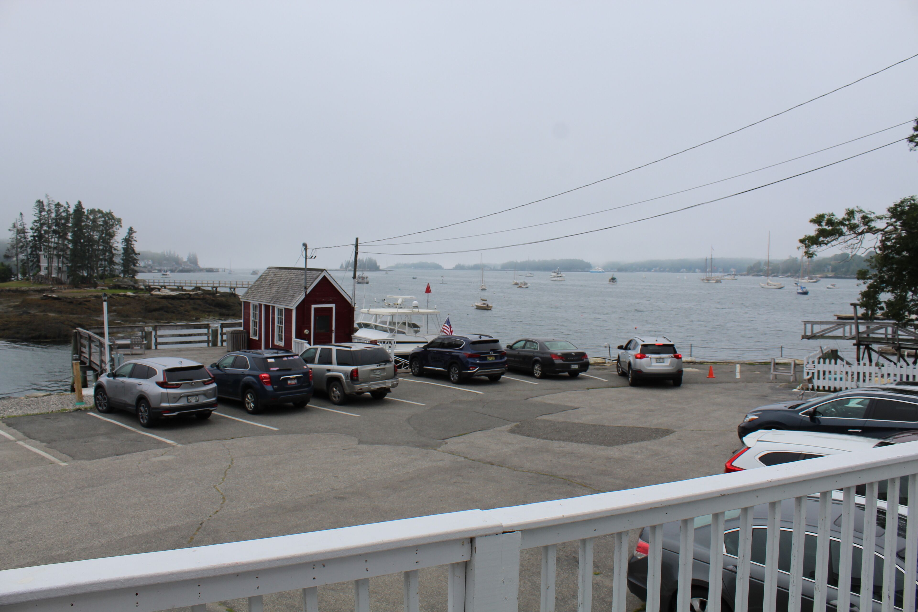 A parking area with several cars, a small red building, and a body of water with boats in the distance.