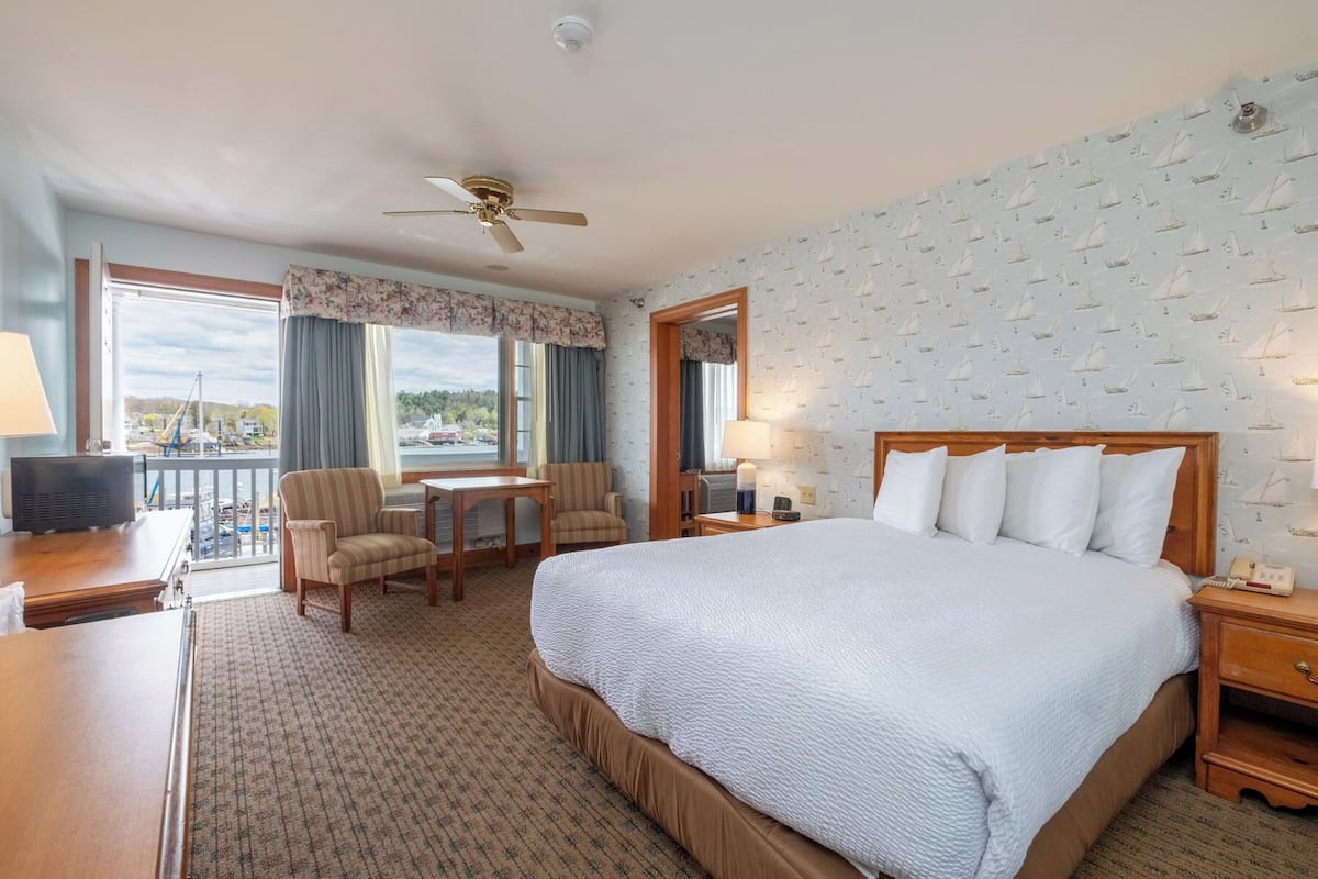 A hotel room with a bed, a desk, a chair, a table, a ceiling fan, and a view of the outside.