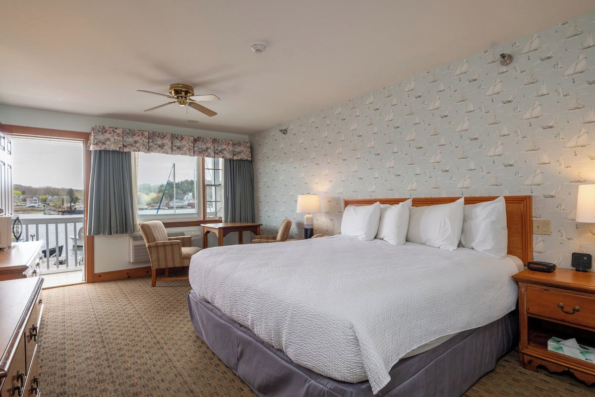 A hotel room with a large bed, a desk, a chair, a balcony with a view, and a ceiling fan.
