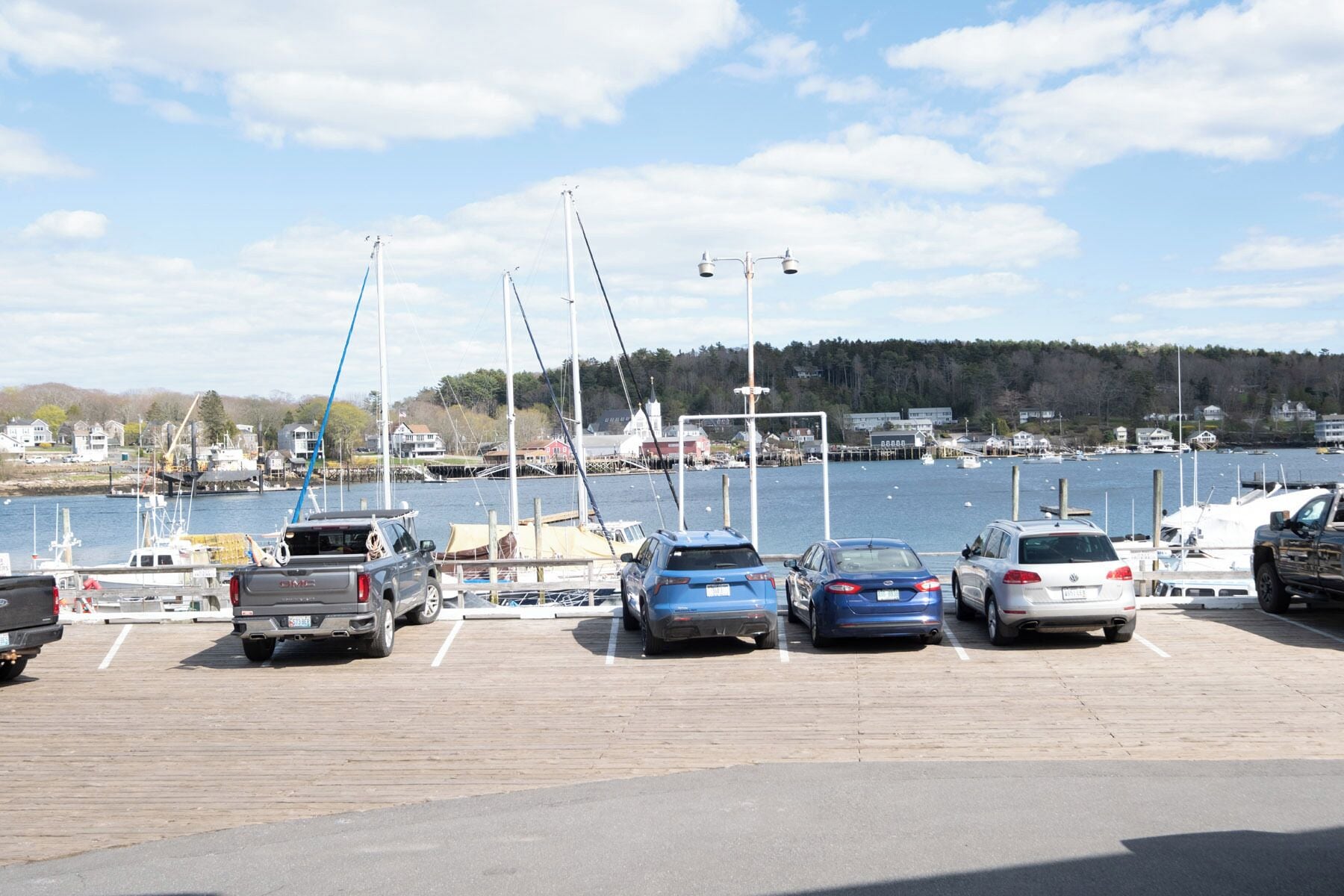 A parking lot with several cars and a truck, a dock with boats, and a body of water with buildings in the background.