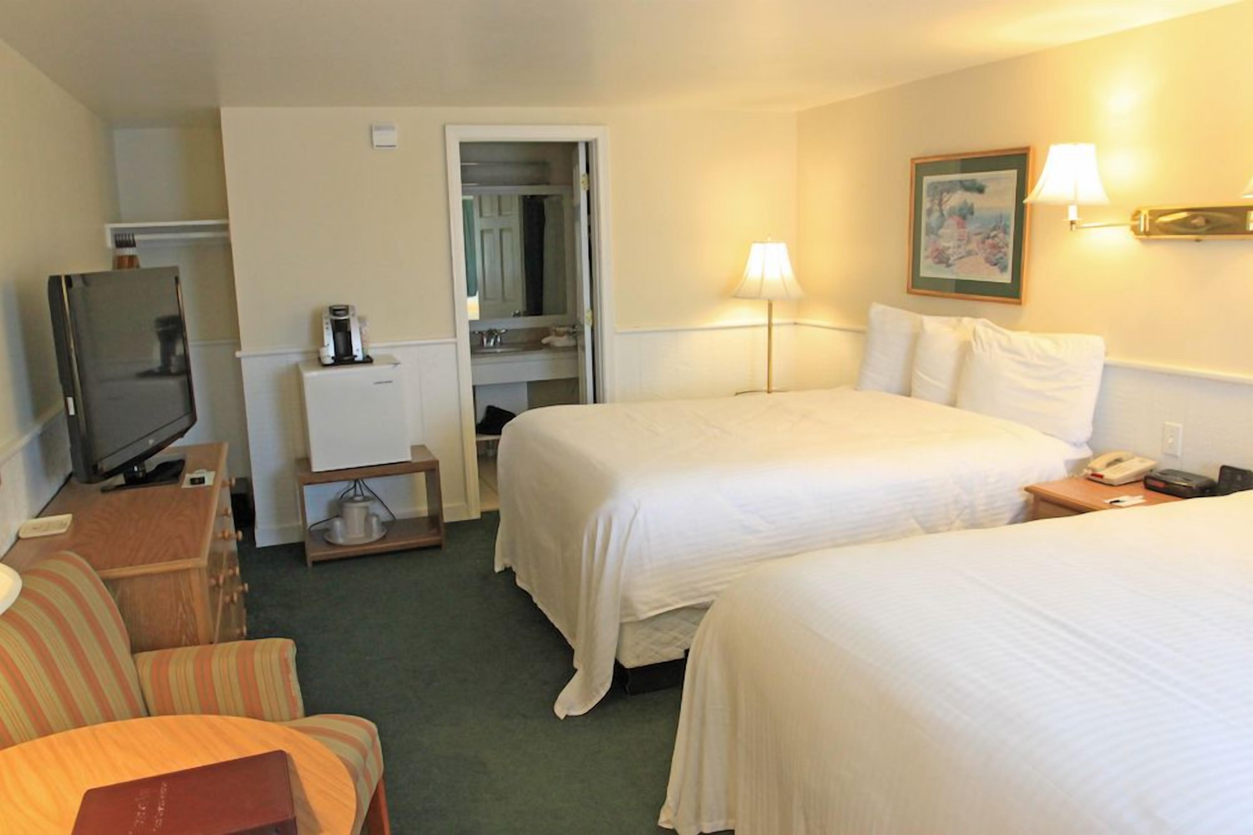 A hotel room with two beds, a television, a desk with a coffee maker, a chair, and a painting on the wall.