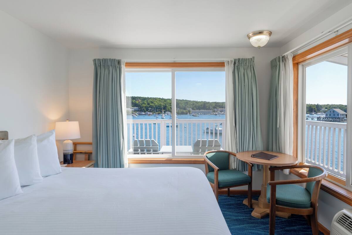 A hotel room with a large bed, a small table with chairs, a view of the sea and boats, and a balcony with a railing.