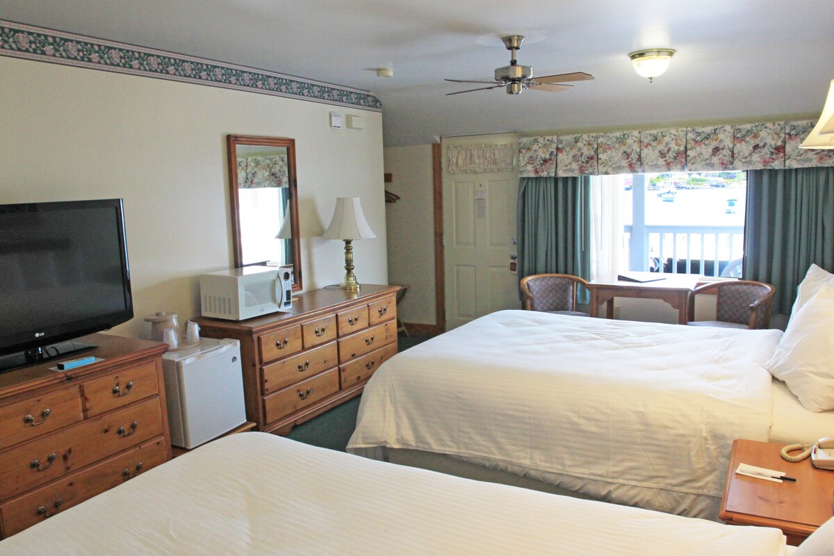A hotel room with two beds, a television, a desk with a chair, a ceiling fan, and a window with curtains.