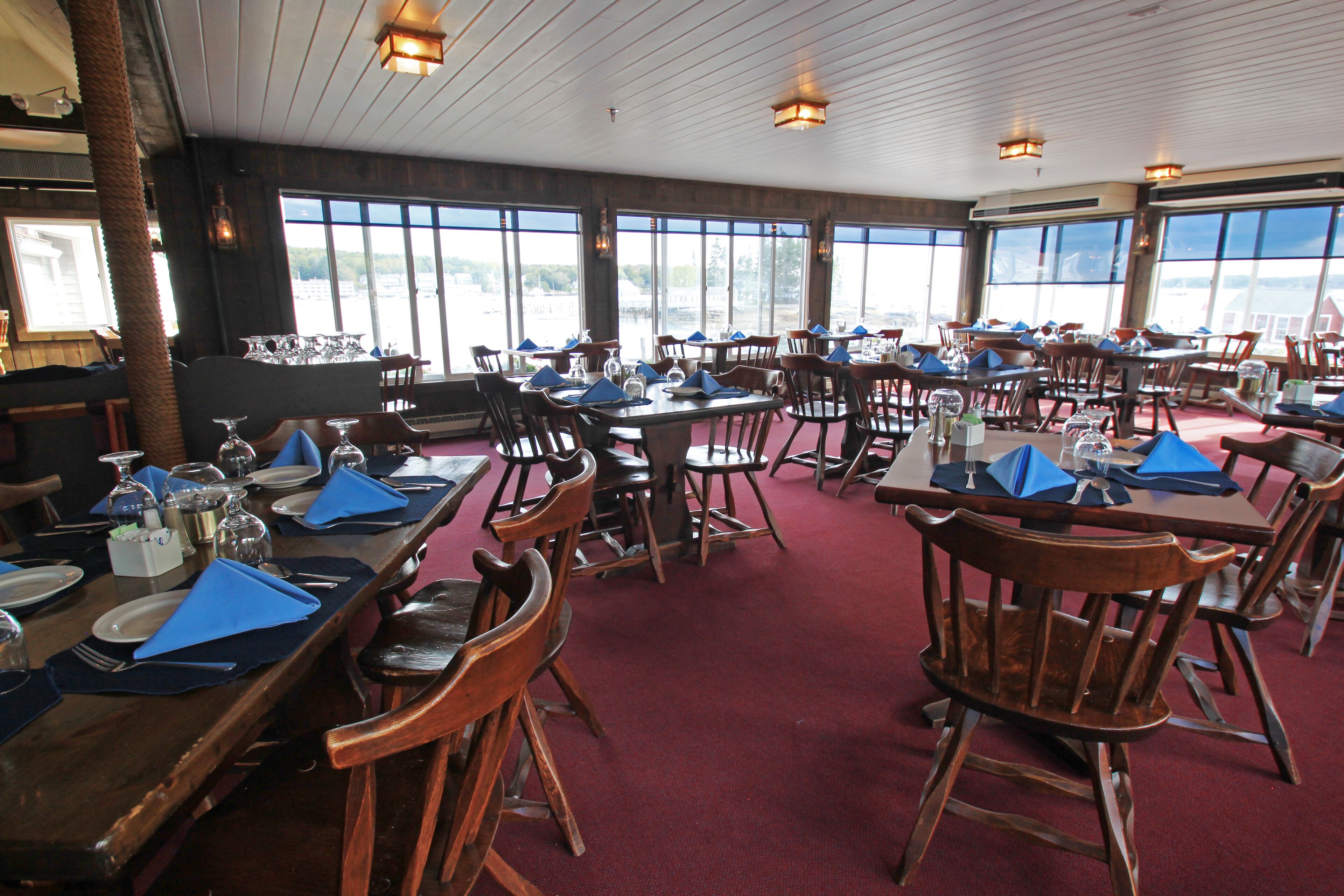 A restaurant with multiple tables and chairs, large windows, and a red carpeted floor.