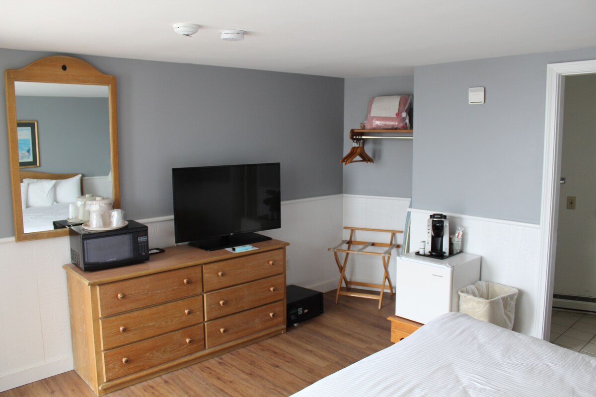 A bedroom with a wooden dresser, a television, a small refrigerator, a chair, and a bed.