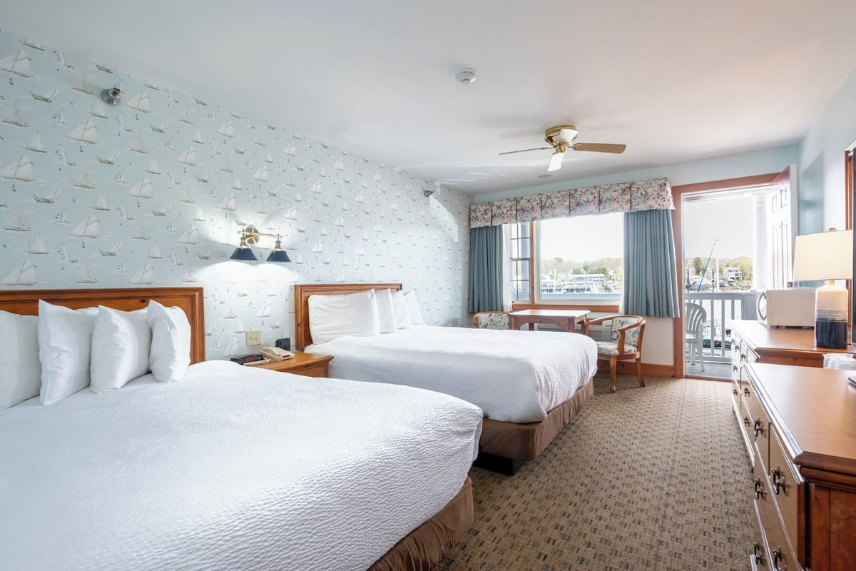 A hotel room with two beds, a desk, a chair, a balcony with a view, and a ceiling fan.