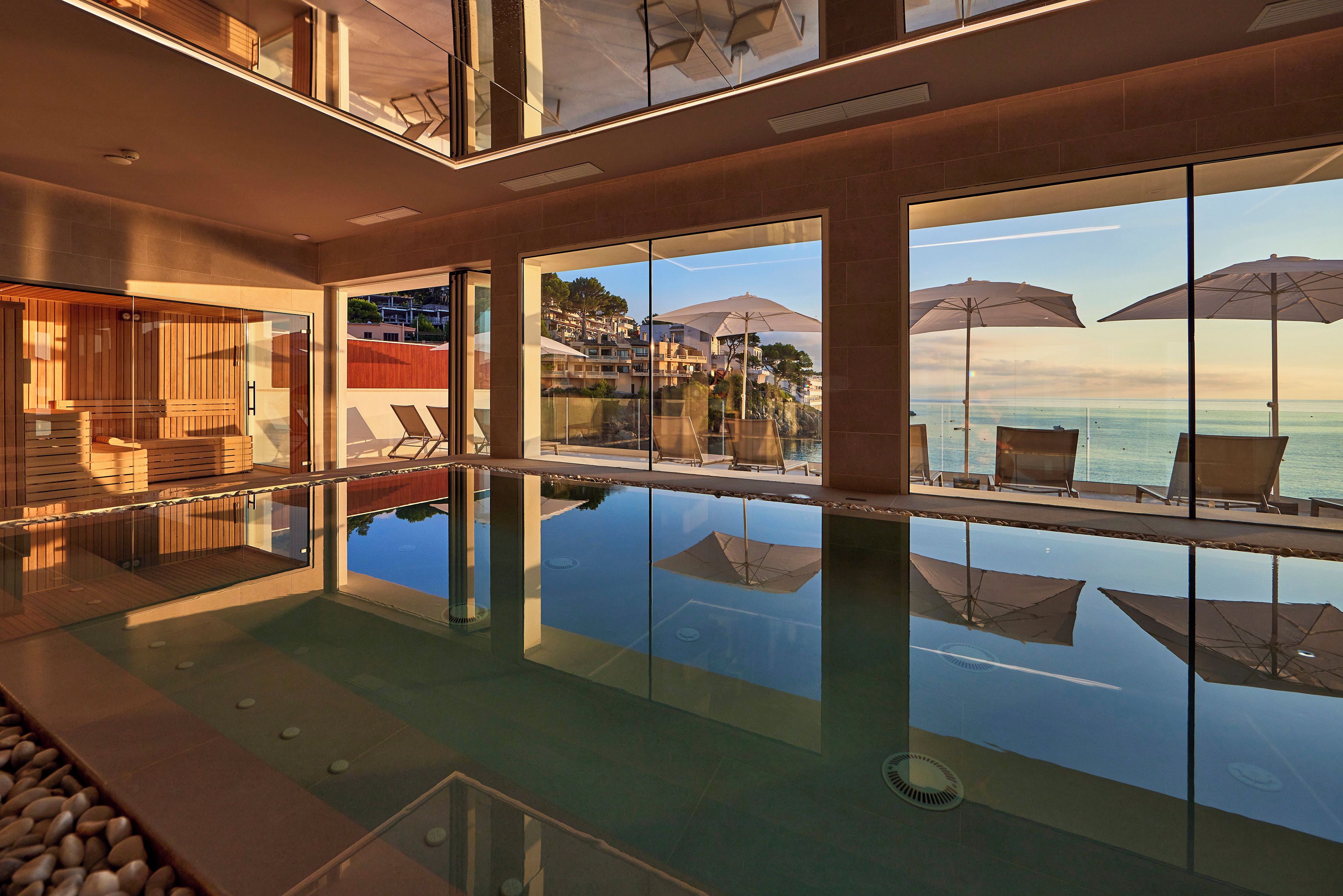 A modern indoor swimming pool with clear glass walls, reflecting the surrounding area, and a view of the sea.