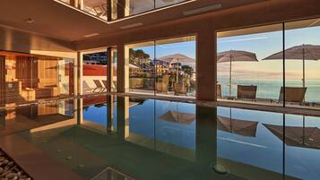 A modern indoor swimming pool with clear glass walls, reflecting the surrounding area, and a view of the sea.