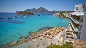 Un hôtel en bord de mer, avec une plage, une eau turquoise cristalline, des bateaux et un paysage montagneux en arrière-plan.