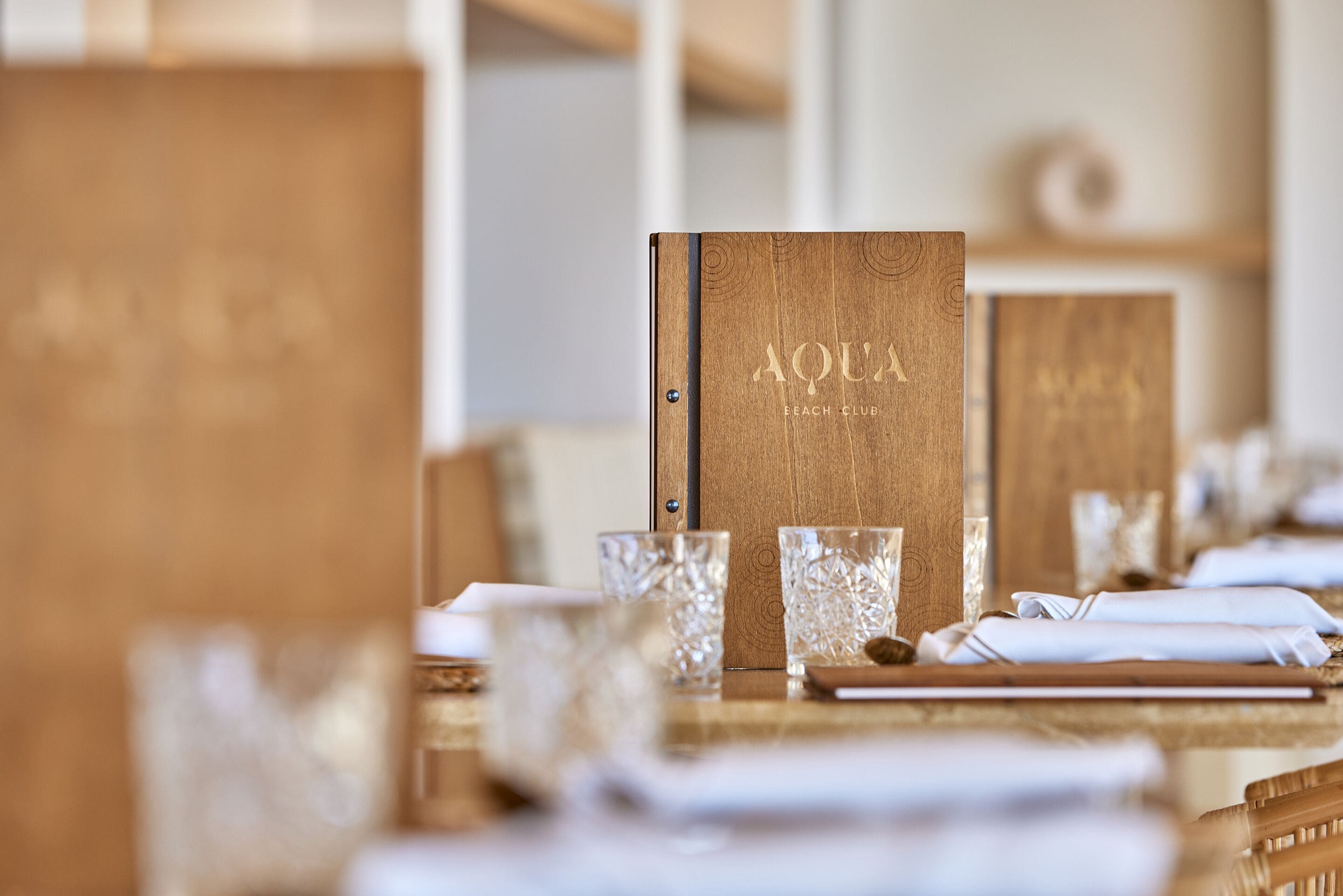 A wooden menu stand labeled "AQUA BEACH CLUB" with a blurred background of a dining area.