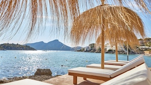 Un salon de plage doté de chaises blanches rembourrées et d’un parasol en paille, offrant une vue sur une mer calme et des montagnes lointaines.