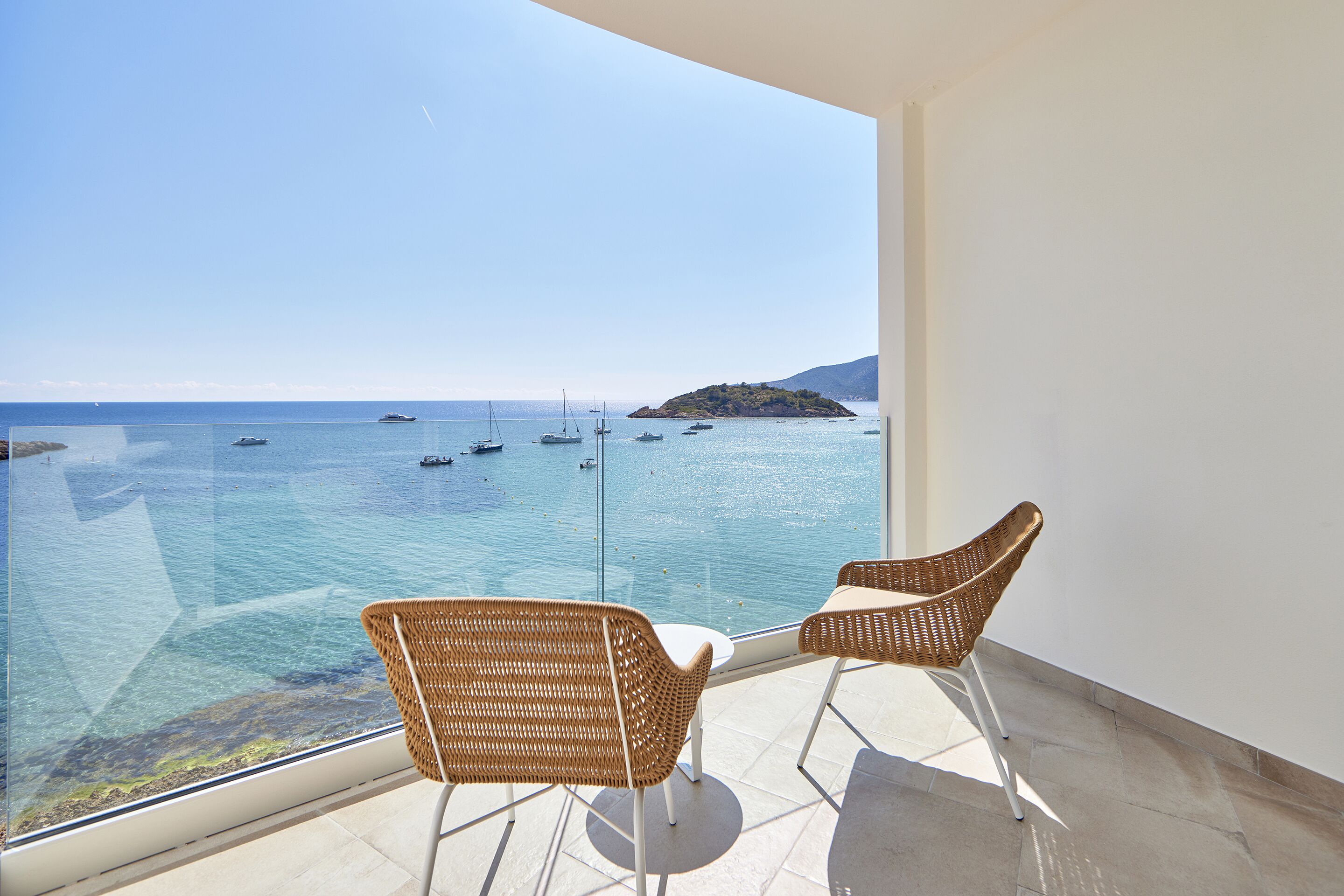 A balcony with wicker chairs and a glass balustrade overlooking a clear blue sea with boats and an island.