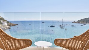 Un balcon agrémenté de fauteuils en osier et d’une petite table, d’où l’on peut admirer une mer bleue et limpide où naviguent plusieurs bateaux.