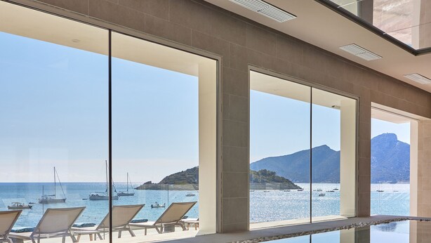 Une piscine intérieure moderne avec des parois en verre transparent, offrant une vue sur les bateaux et un paysage montagneux.