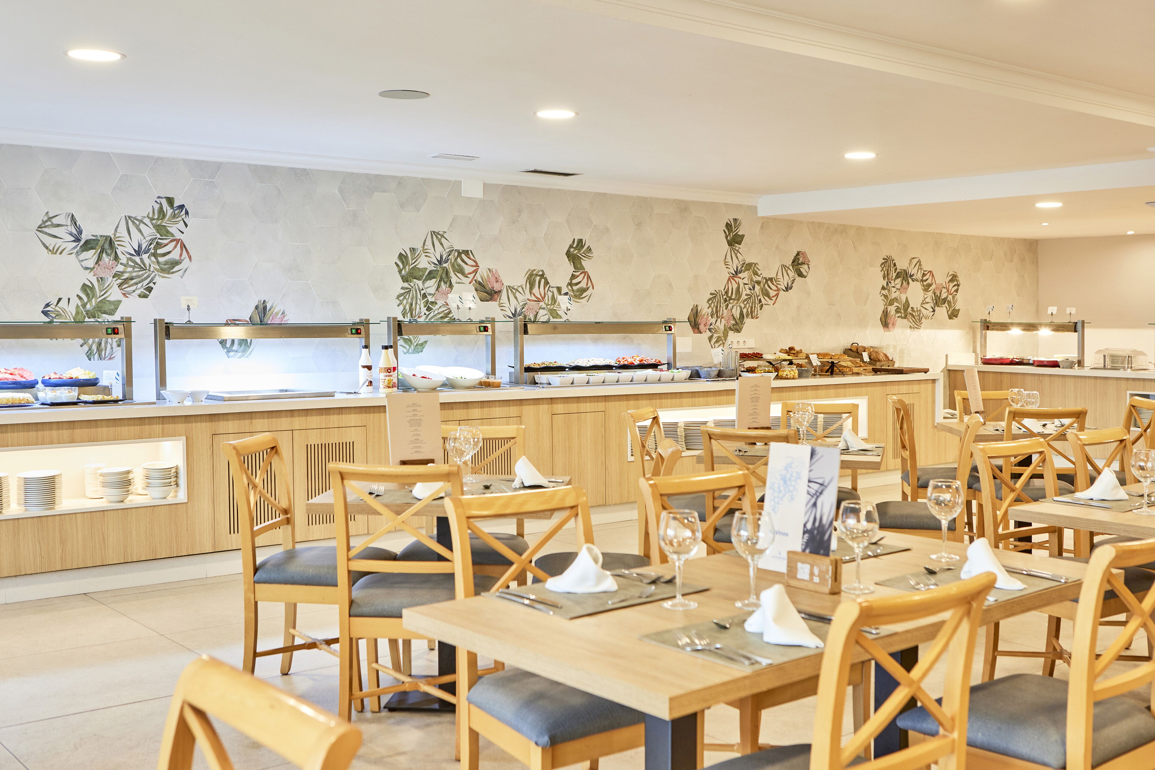 Un restaurante moderno con mesas y sillas de madera, una zona de bufé y una pared decorada con motivos de hojas tropicales.