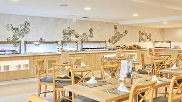 Un restaurante moderno con mesas y sillas de madera, una zona de bufé y una pared decorada con motivos de hojas tropicales.