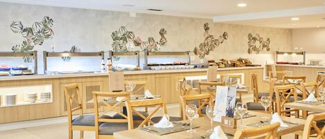 Um restaurante moderno com mesas e cadeiras de madeira, área de buffet e parede decorada com motivos de folhas tropicais.
