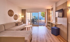 A modern hotel room with a sofa, a small table, a TV mounted on the wall, and a view of the sea through the glass doors. - Universal Aparthotel Don Camilo (Andraitx)