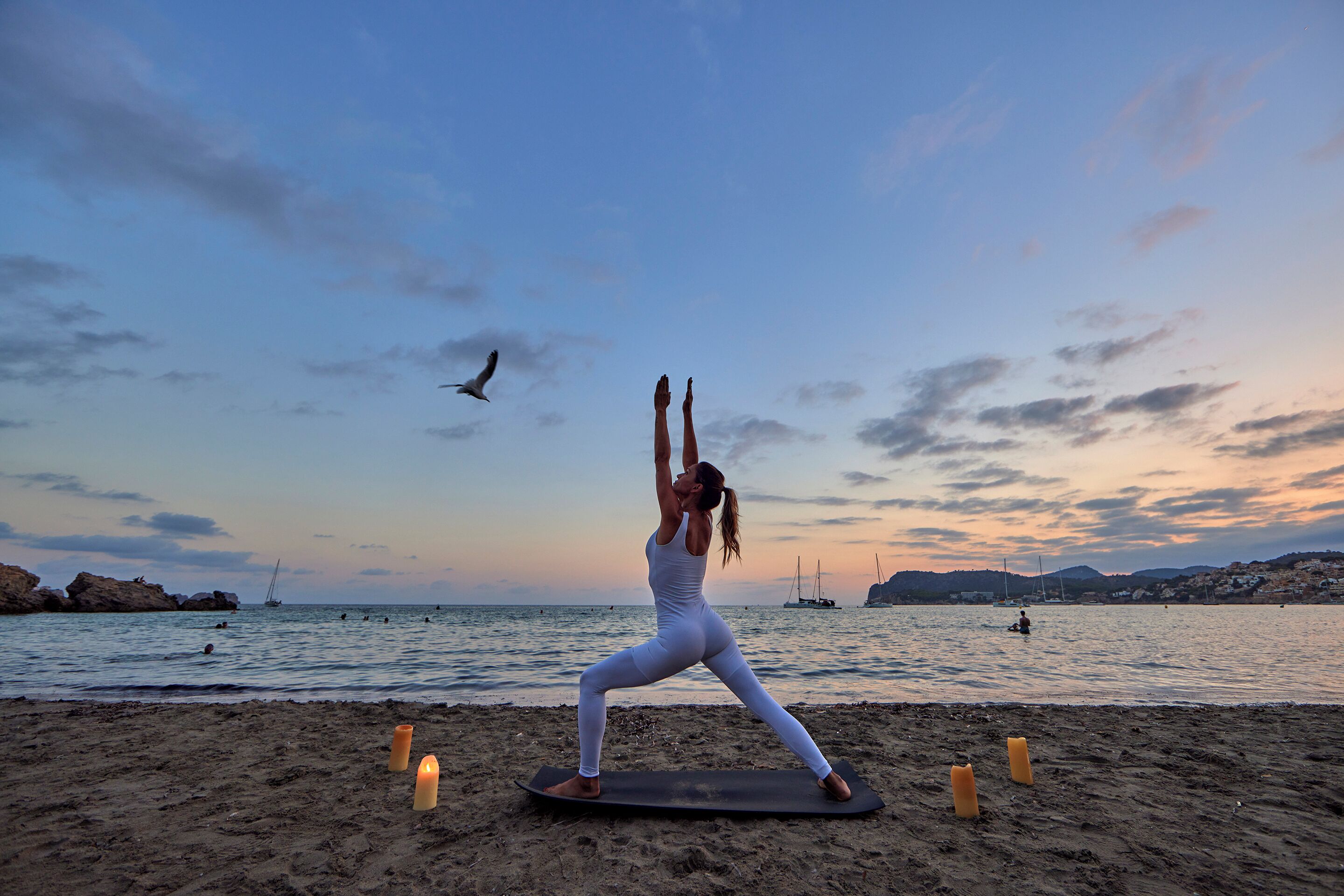 Une personne pratiquant le yoga sur un tapis, sur une plage, avec une mer calme et des bateaux en arrière-plan.