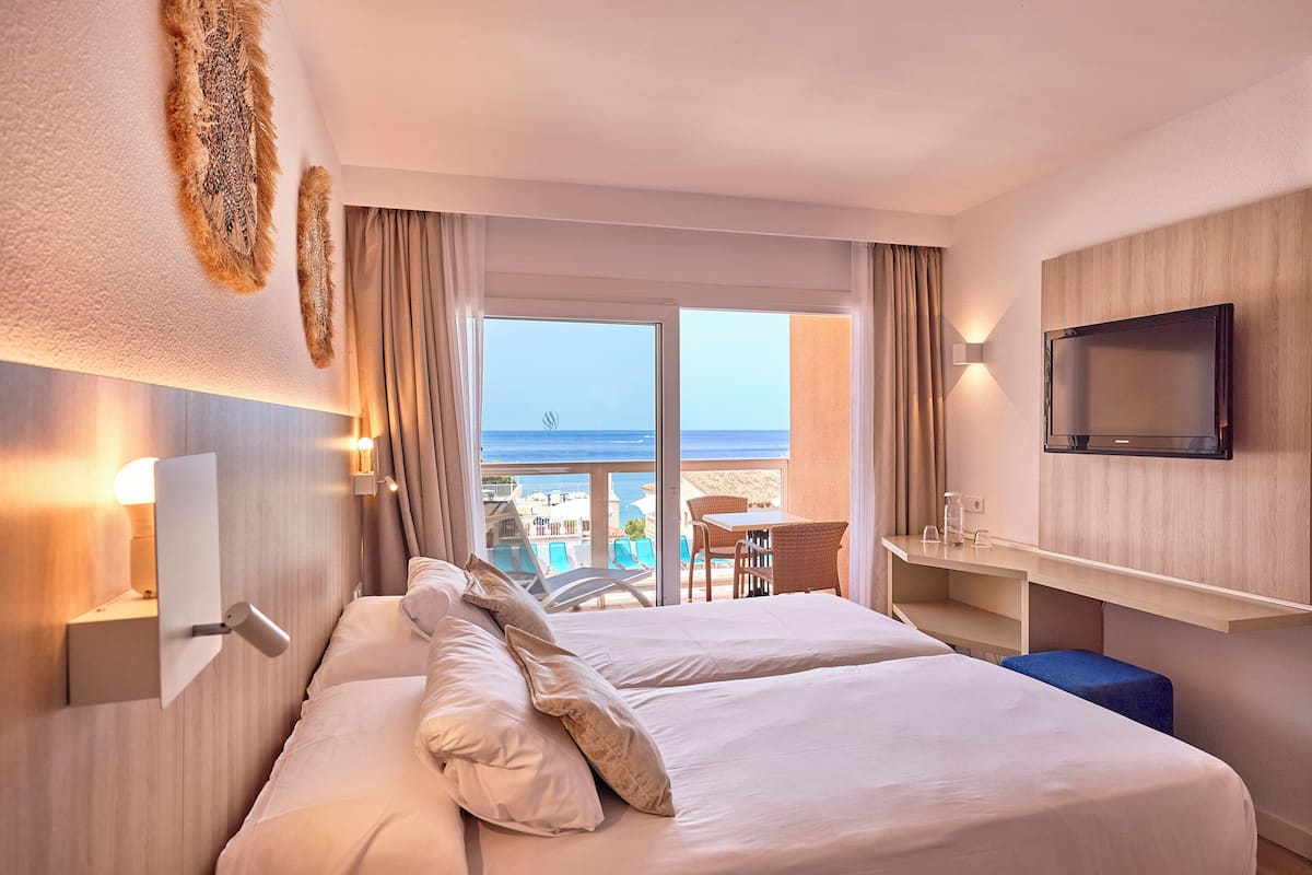 A hotel room with two beds, a TV, a desk, and a balcony with a view of the sea.