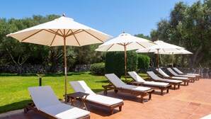 A row of wooden lounge chairs with white cushions and beige umbrellas, arranged on a tiled patio with a stone wall and greenery in the background.